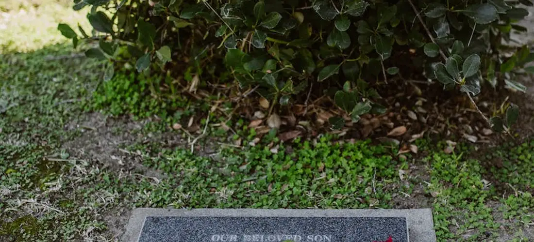 A low grave marker on the ground surrounded by grass and shrubbery, conveying a quiet funeral setting.