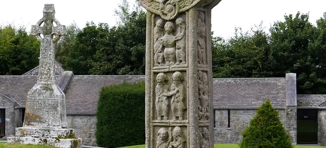 Cemetery scene with tall carved stone monuments and a cross-shaped headstone, a church-like building in the background, and green shrubs.