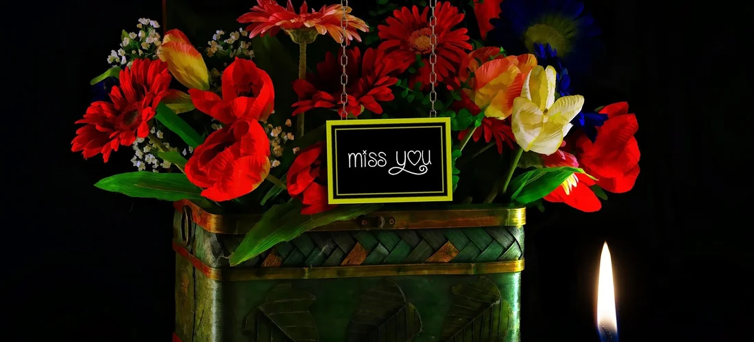 A bright bouquet of red, orange, and yellow flowers in a green container with a small chalkboard sign that reads 'miss you' and a lit candle nearby.