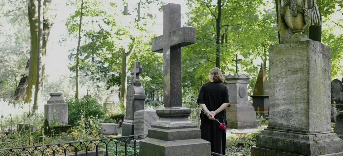 A person stands with hands behind their back in a cemetery, surrounded by gravestones and greenery.