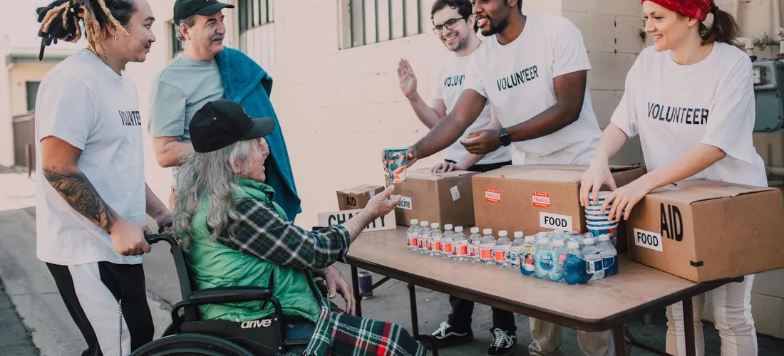 Volunteers in white VOLUNTEER shirts hand a box of supplies to a person in a wheelchair at a donation event.