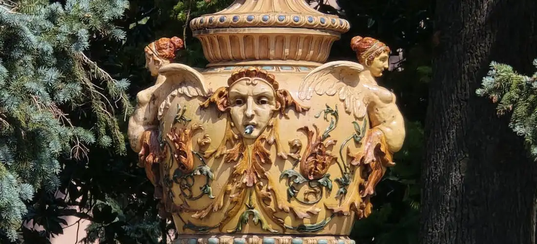 Ornate ceramic urn with carved faces and figures in a garden setting