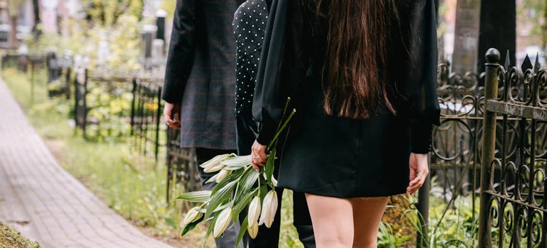 Two mourners walk along a cemetery path; one person holds a bouquet of white flowers as they walk away.