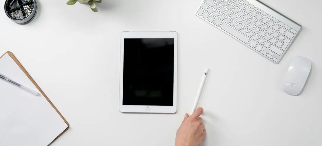 A tidy workspace with a tablet, keyboard, and a hand holding a pencil at a white desk, symbolizing careful writing.