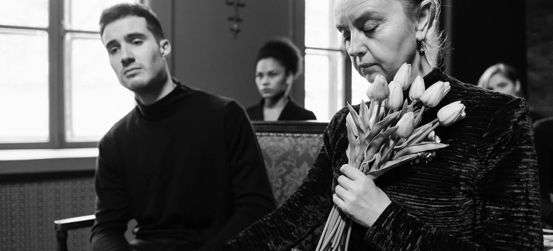 A somber, non-religious funeral scene with attendees seated; a person on the right holds a bouquet of flowers while others sit in quiet contemplation.