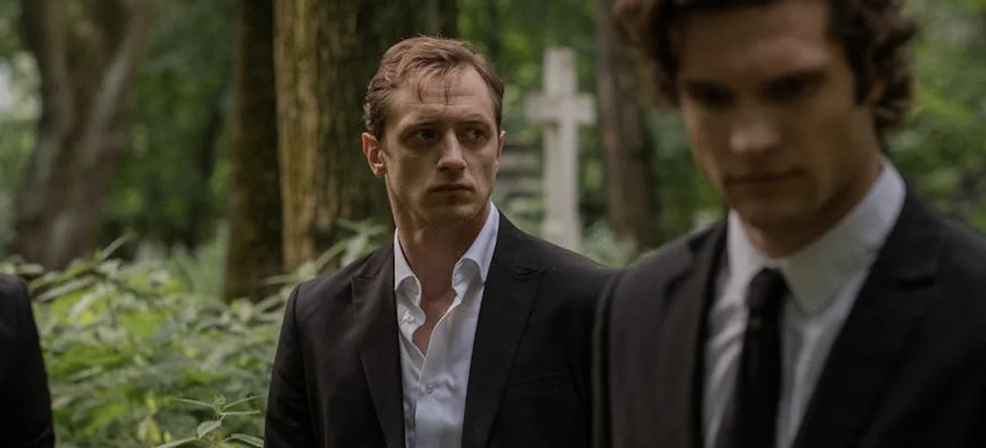 Two men in dark suits stand in a wooded cemetery with blurred grave markers in the background, conveying a somber moment.
