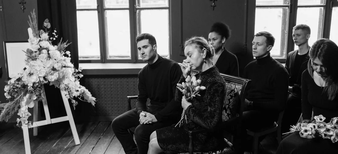 Black-and-white photo of mourners seated in a funeral service, with a large floral arrangement nearby and light coming through windows.