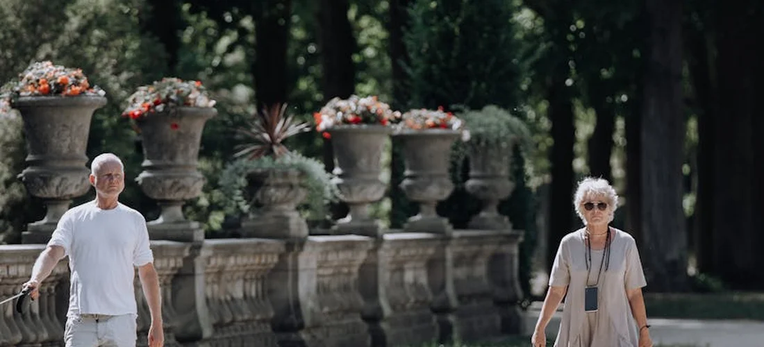 Two adults walk along a garden path between tall stone planters filled with flowers.