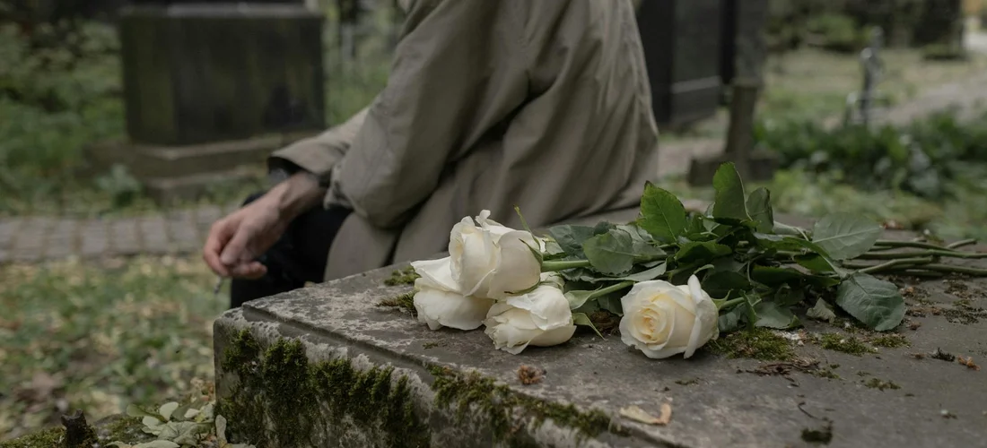 A cemetery scene with a tombstone and white roses laid on its edge, while a person in a coat stands nearby.