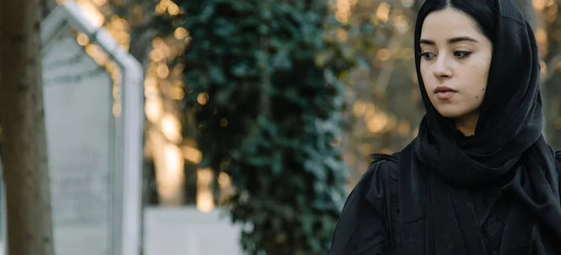 A solemn woman in a black hijab stands outdoors, eyes lowered in contemplation.