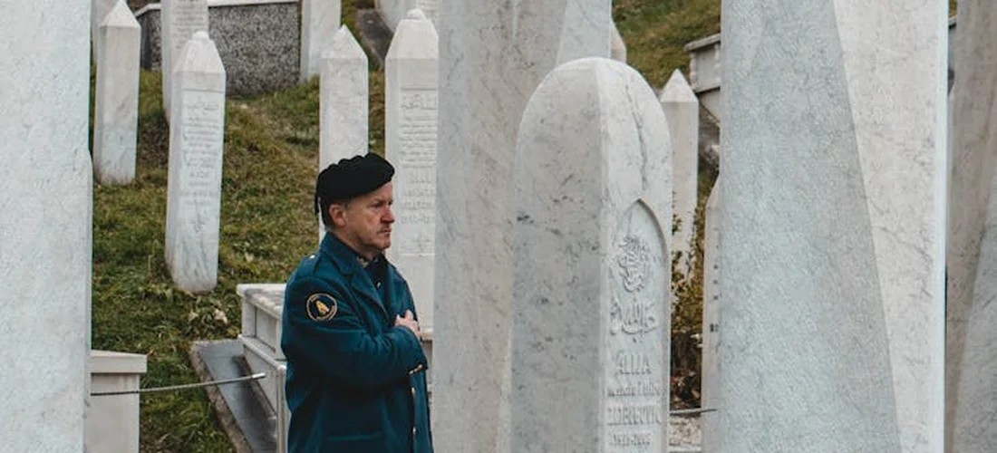 A person in a dark jacket stands among tall white gravestones in a cemetery, hands pressed together near the chest, looking contemplative.