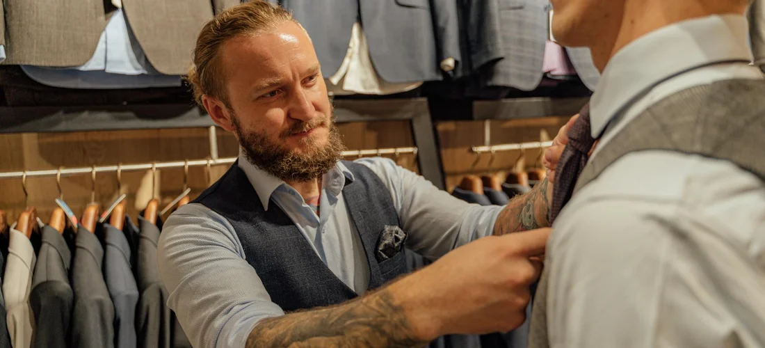 A man adjusts a tie on another man in a formal clothing store, with suits and jackets on display in the background.
