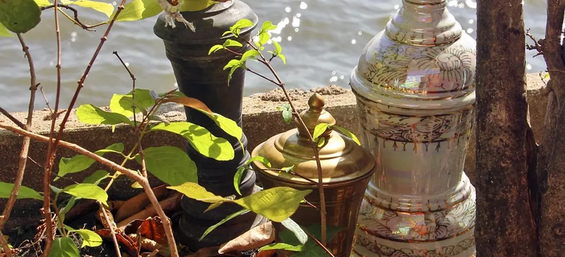 Decorative metal cremation urns partially obscured by green leaves beside a calm body of water.